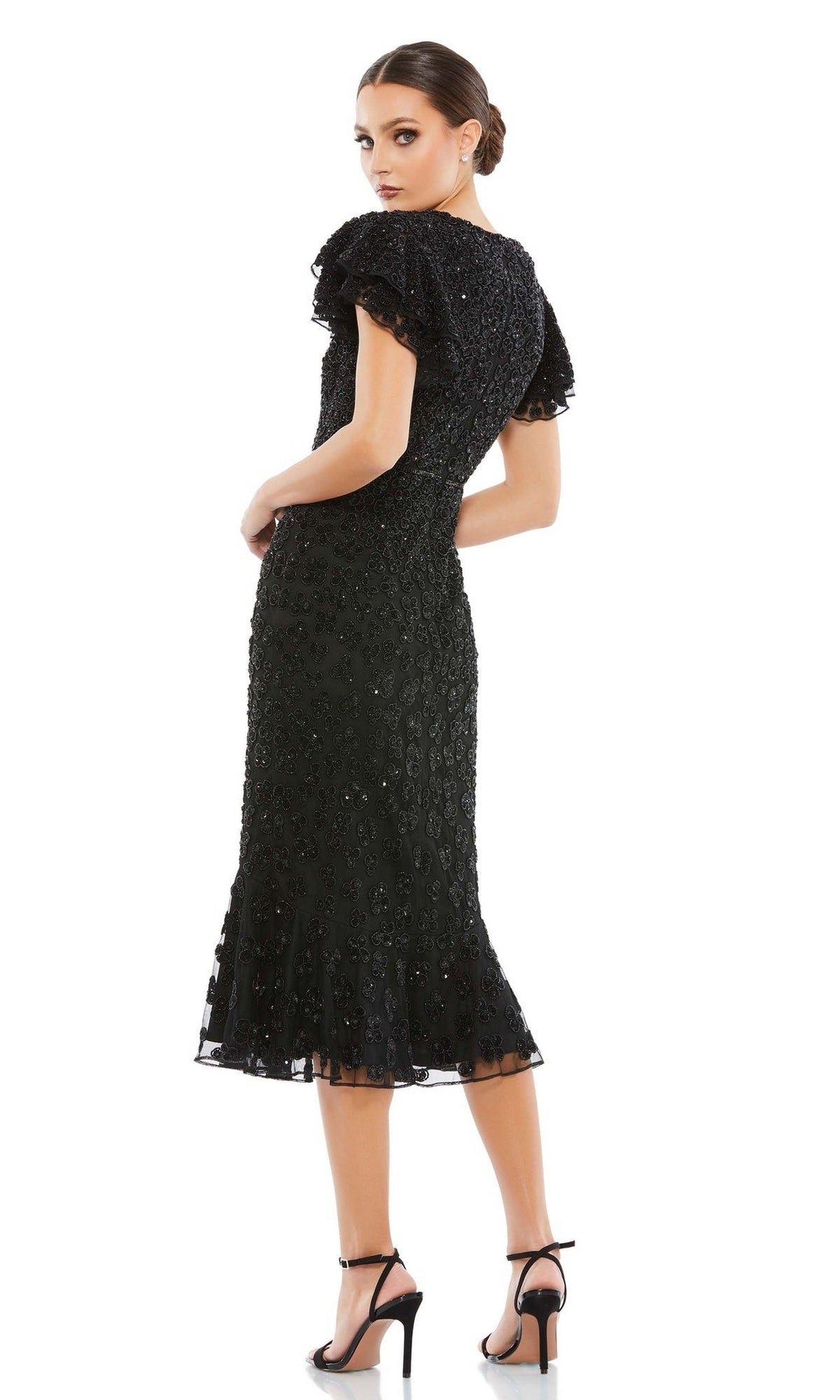 Woman wearing the Short Wedding Guest Dress 10771 by Mac Duggal, a fitted midi black beaded dress with a ruffle hem, looks over her shoulder and pairs it with black high-heeled sandals against a white background.